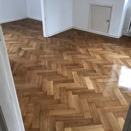 M&L Parkiet - Parkiet ułożony w jodełkę, świeżo po lakierowaniu, z widocznym połyskiem. Ujęcie z perspektywy wejścia do pomieszczenia, widoczne drzwi i fragment ściany.