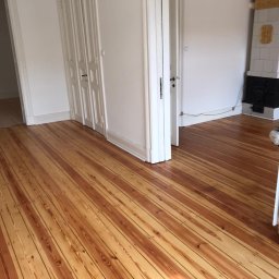 M&L Parkiet - Wnętrze po cyklinowaniu: parkiet z widocznym usłojeniem, białe ściany, drzwi i piec kaflowy w tle. Jasne, naturalne światło podkreśla odnowioną podłogę.