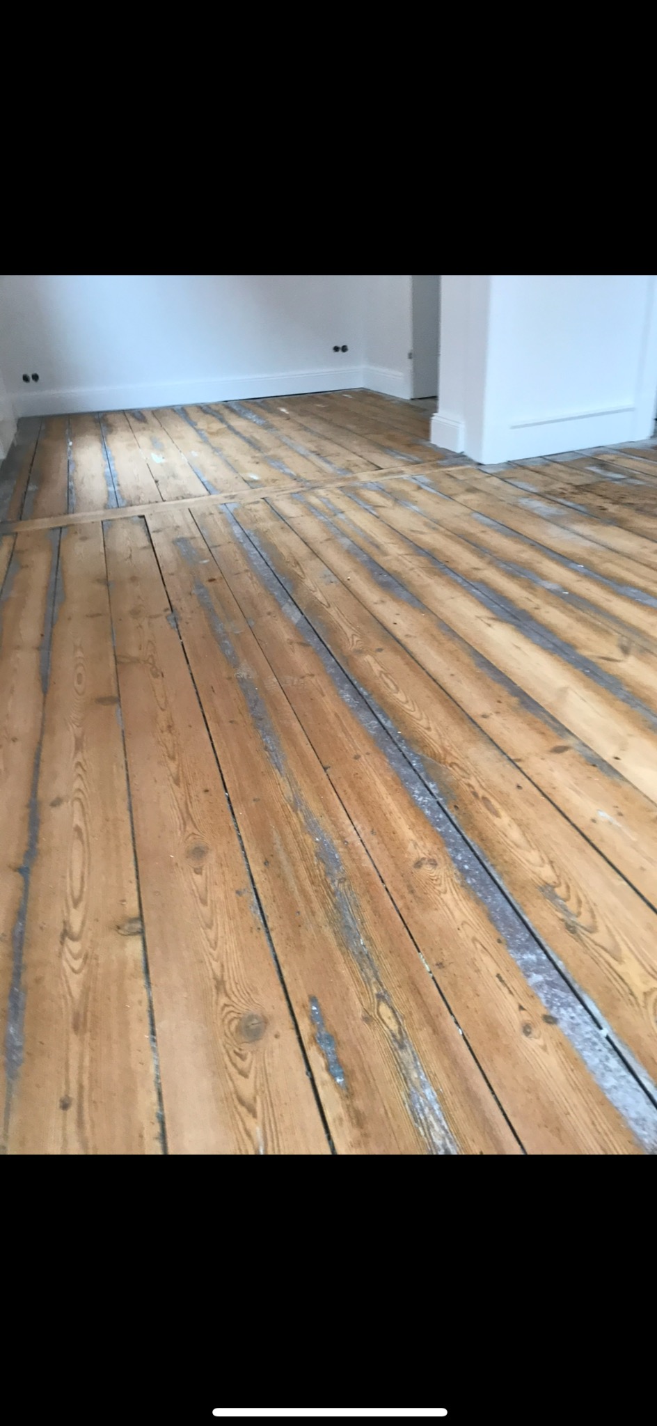 Drewniana podłoga w trakcie renowacji, widoczne ślady po szlifowaniu i ubytki, jasne ściany w tle. Podłoga z wyraźnym usłojeniem, przygotowana do dalszej obróbki.