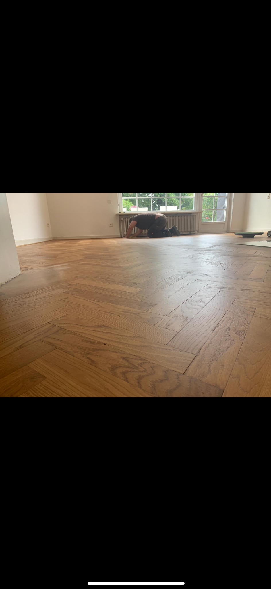 Parkiet ułożony w jodełkę, świeżo po renowacji. Widoczny pracownik w trakcie wykańczania podłogi przy oknie. Jasne wnętrze, naturalne światło.