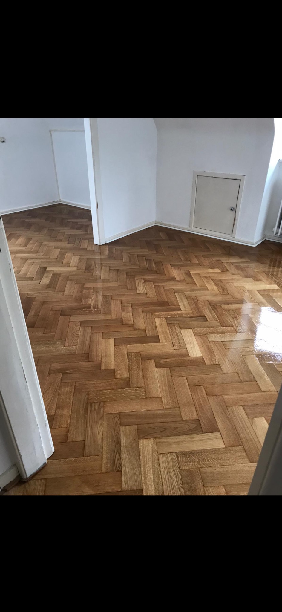 Parkiet ułożony w jodełkę, świeżo po lakierowaniu, z widocznym połyskiem. Ujęcie z perspektywy wejścia do pomieszczenia, widoczne drzwi i fragment ściany.