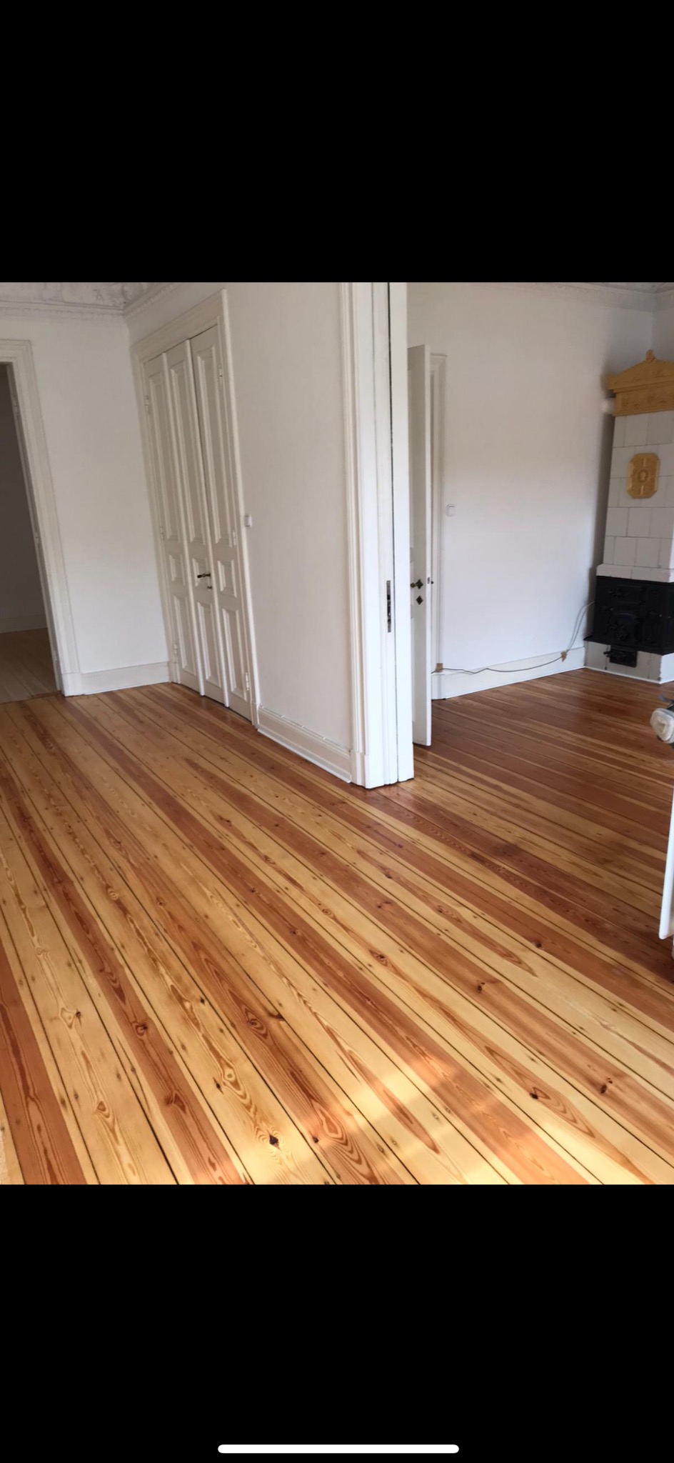 Wnętrze po cyklinowaniu: parkiet z widocznym usłojeniem, białe ściany, drzwi i piec kaflowy w tle. Jasne, naturalne światło podkreśla odnowioną podłogę.