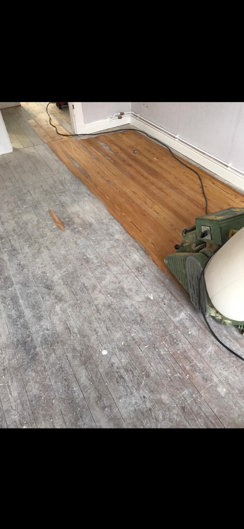 Fragment podłogi z desek: część po cyklinowaniu, ukazująca naturalne drewno, a część przed, z szarą, zniszczoną powierzchnią. Widoczna maszyna do cyklinowania.