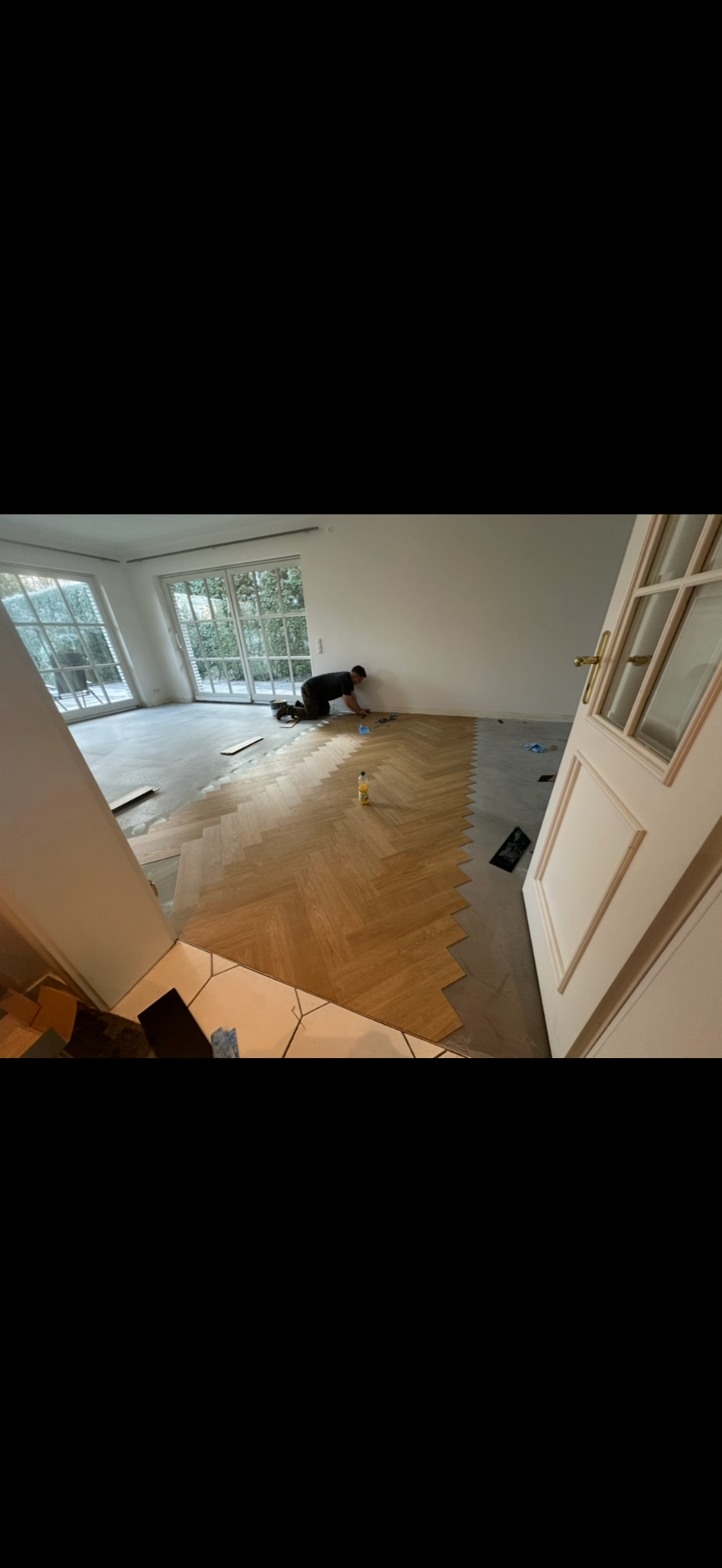 Układanie parkietu w jodełkę w jasnym salonie. Widoczny fragment ułożonej podłogi i pracownik na kolanach, dopasowujący kolejne elementy. Duże okna w tle.