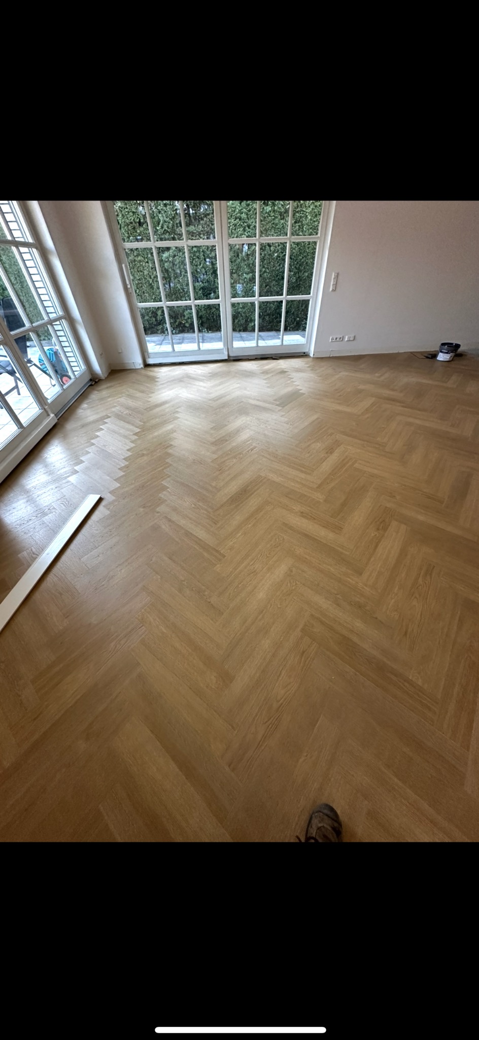 Jasny parkiet ułożony w jodełkę francuską w przestronnym pokoju z dużymi oknami balkonowymi. Widoczny fragment listwy przypodłogowej i but.