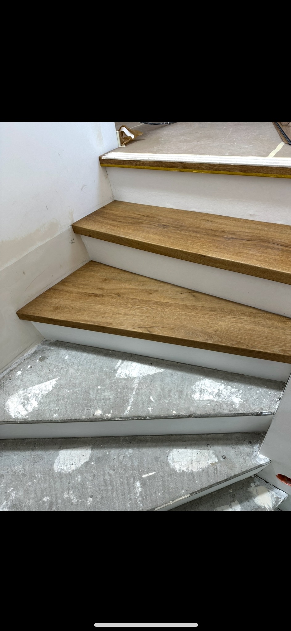 Dwa stopnie schodów z drewnianymi nakładkami, białe boki, surowy beton na dolnym stopniu. Widok z góry, ukośnie. Detal wykończenia schodów.