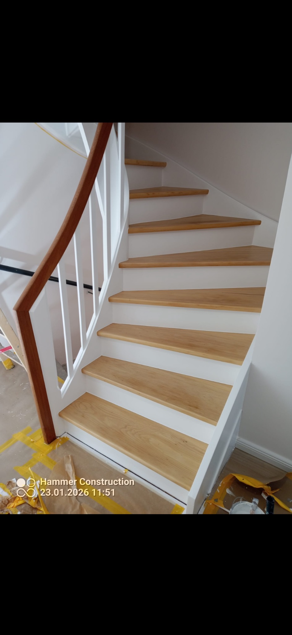 Jasne, drewniane schody z białymi podstopnicami i brązową poręczą. Widoczne zabezpieczenia podłogi. Ujęcie z dołu, podkreślające detale wykończenia.