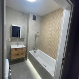 SK&A-Technika Serhii Kushnir - Nowoczesna łazienka z podświetlaną wanną, drewnianą ścianą i szafką pod umywalką. Szare płytki i okrągła lampa sufitowa tworzą spójny design.