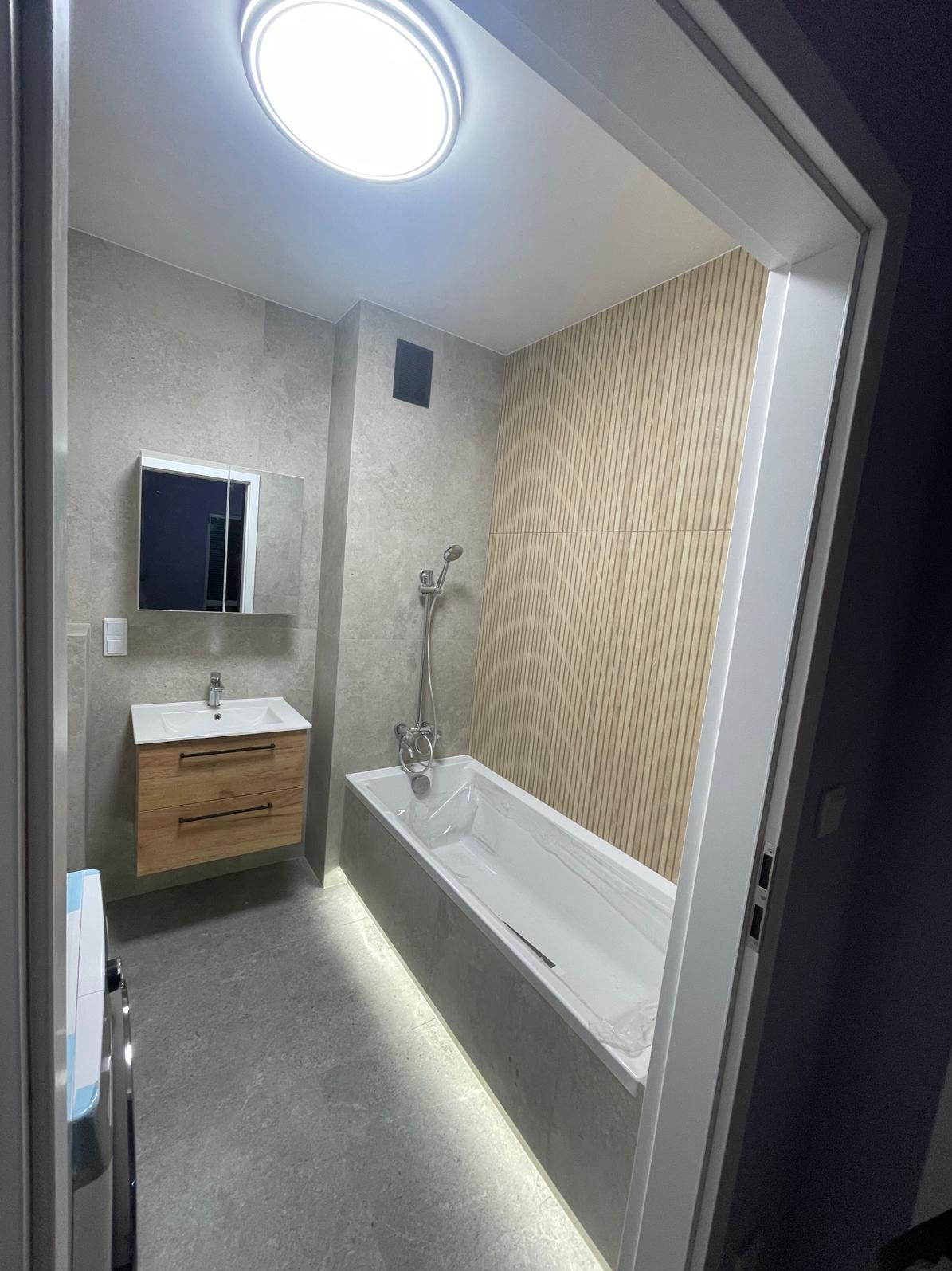 Nowoczesna łazienka z podświetlaną wanną, drewnianą ścianą i szafką pod umywalką. Szare płytki i okrągła lampa sufitowa tworzą spójny design.