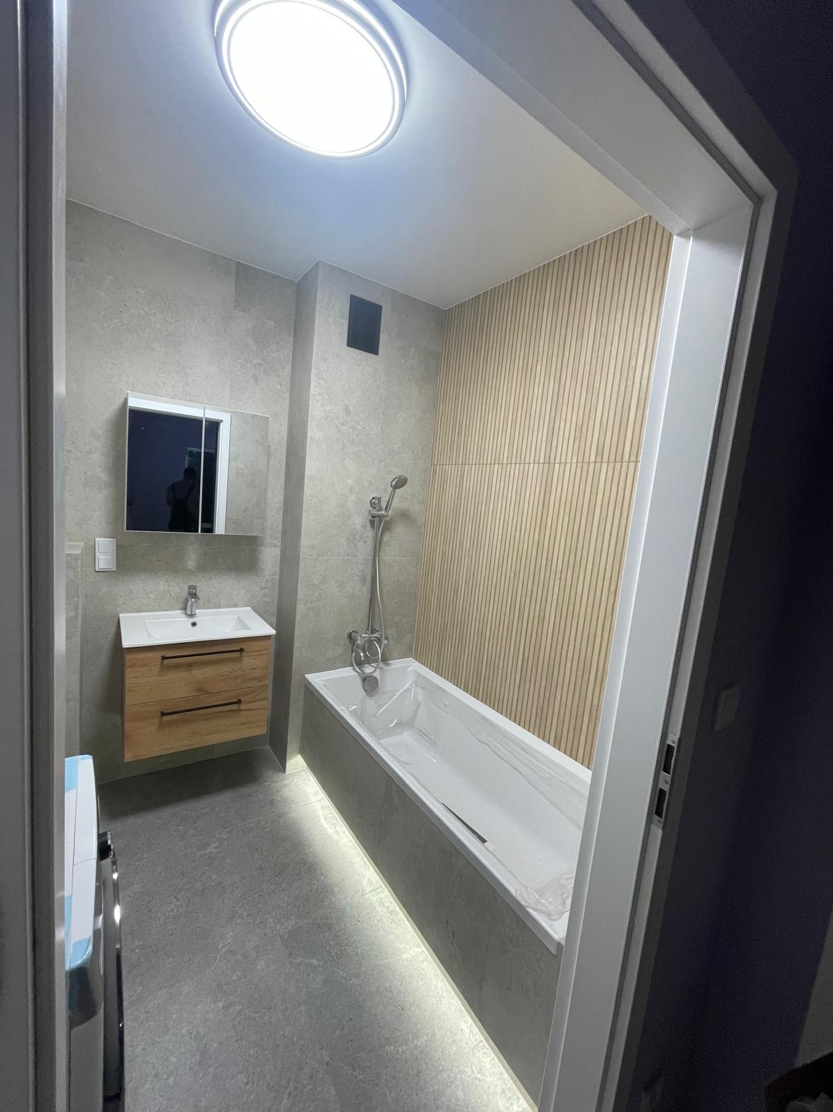 Nowoczesna łazienka z podświetlaną wanną, drewnianą szafką pod umywalką i panelami ściennymi. Minimalistyczny design z szarymi płytkami i okrągłym oświetleniem sufitowym.