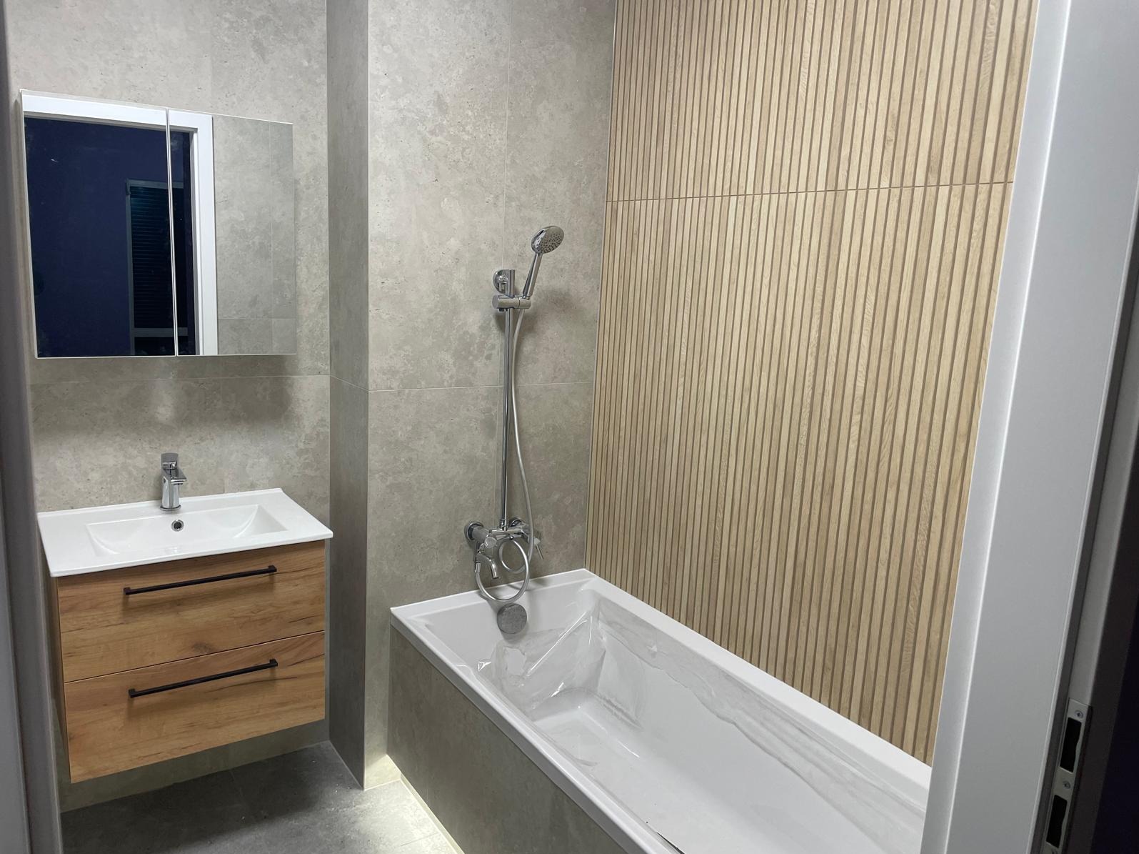 Wykończona łazienka w Poznaniu: drewnopodobna ściana, wanna z baterią, umywalka z szafką i lustro. Nowoczesny design, szare płytki, minimalistyczny styl.