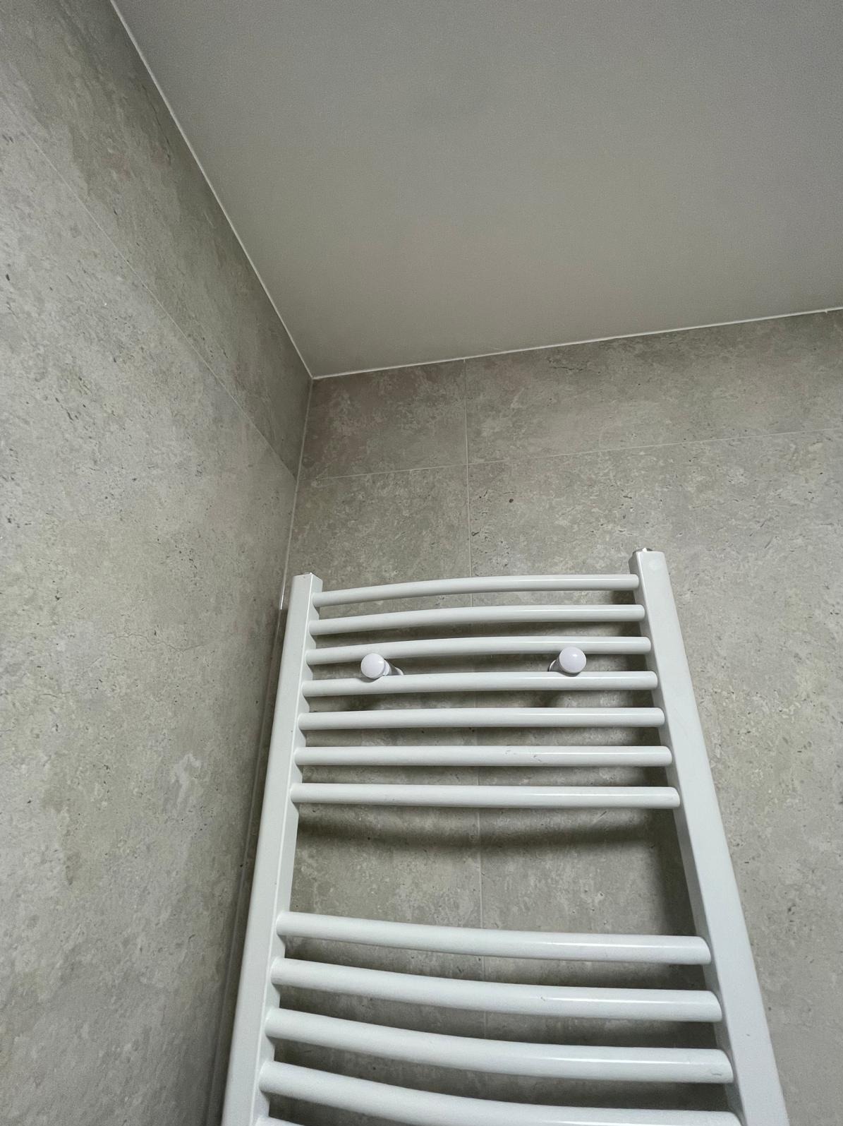 Biały grzejnik drabinkowy z dwoma haczykami na tle szarych płytek w łazience. Ujęcie z dołu, widoczny fragment sufitu.