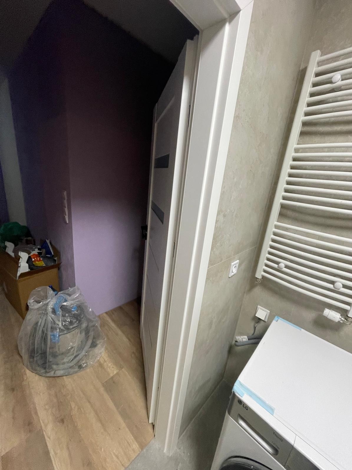 Białe drzwi z ościeżnicą w łazience z grzejnikiem drabinkowym i pralką. Widoczne materiały budowlane i fioletowa ściana w tle. Ukończony montaż.