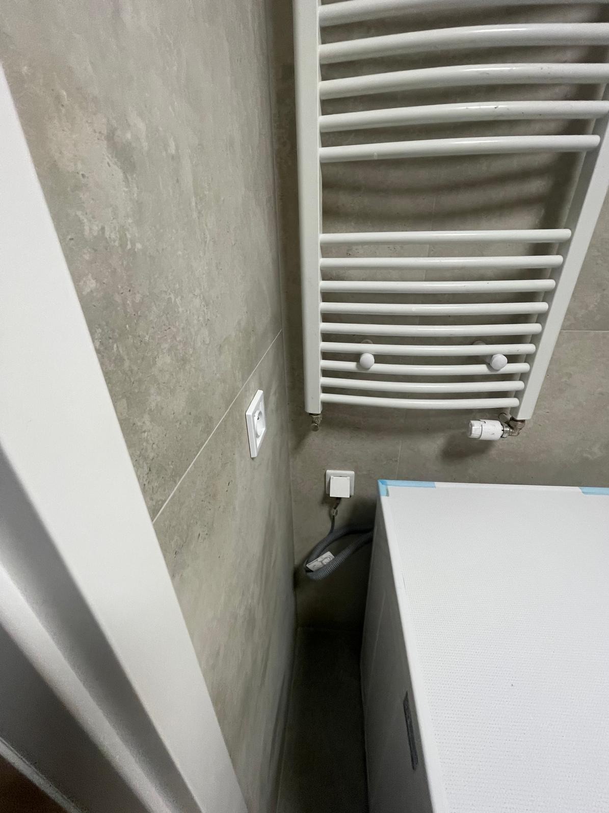 Szary gres w łazience z białym grzejnikiem drabinkowym, gniazdkiem i fragmentem pralki. Minimalistyczny design, neutralna kolorystyka, widok z perspektywy.