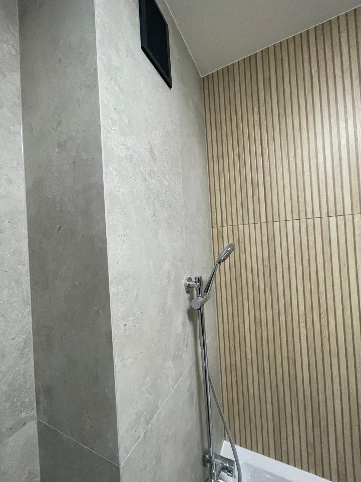 Nowoczesna łazienka z szarymi płytkami imitującymi kamień i drewnianą ścianą z pionowymi panelami. Widoczna bateria prysznicowa i kratka wentylacyjna.