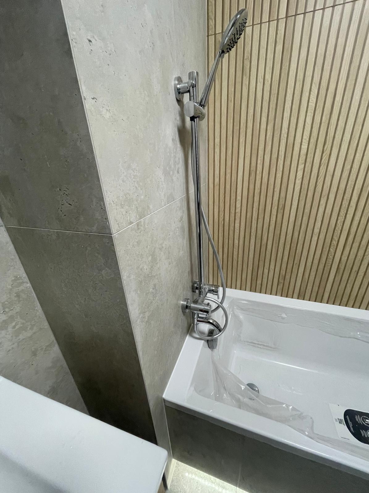 Nowoczesna łazienka: chromowana bateria natryskowa przy białej wannie, ściany wyłożone płytkami imitującymi beton i drewno. Minimalistyczny design, stonowana kolorystyka.