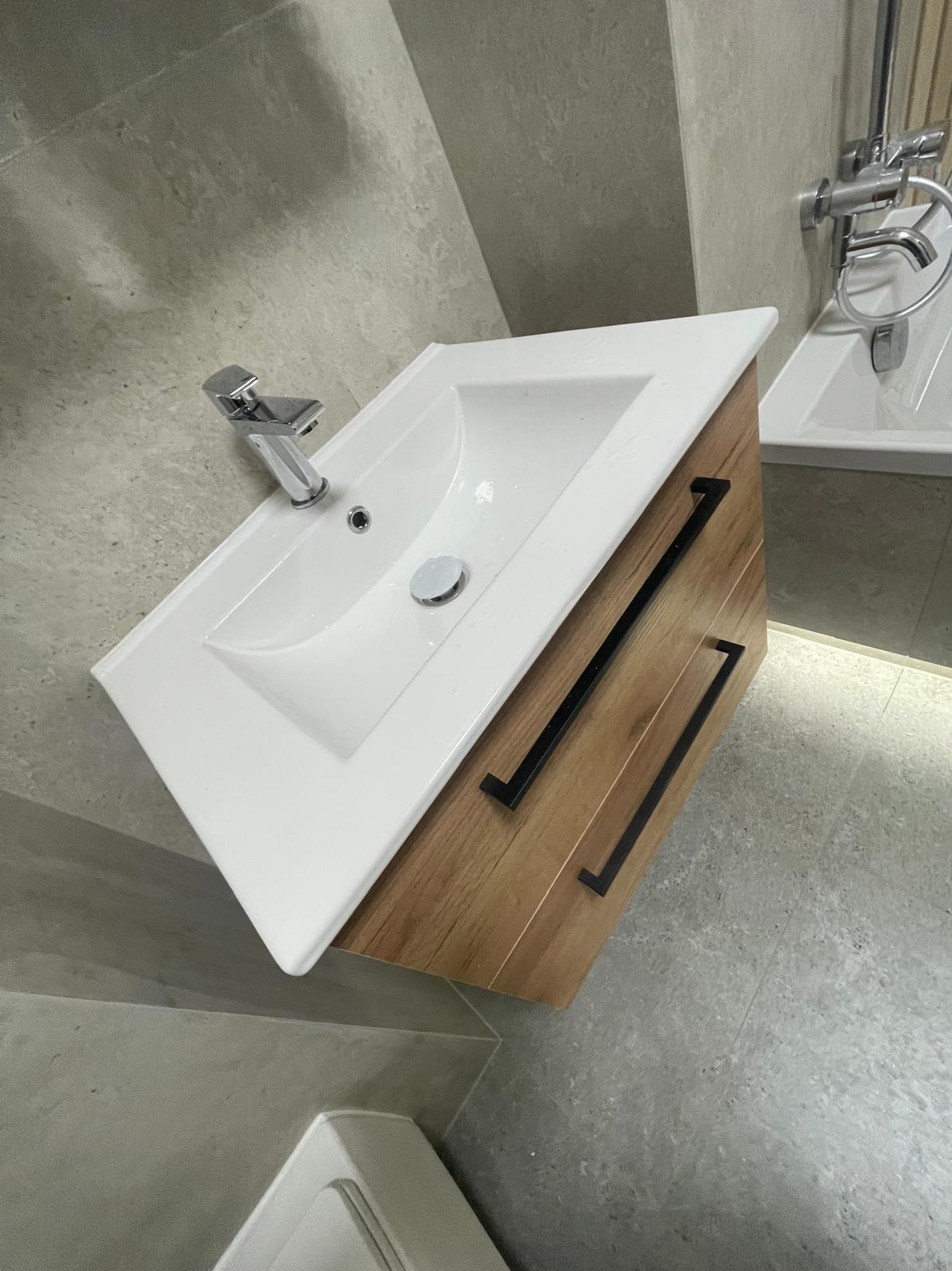 Nowoczesna biała umywalka z drewnianą szafką i czarnymi uchwytami w łazience wykończonej szarymi płytkami. Widoczna bateria i fragment wanny.