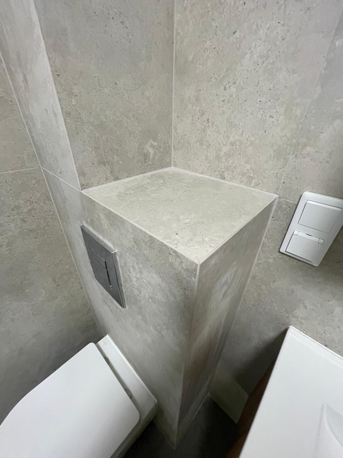 Minimalistyczna łazienka z zabudową WC w odcieniach szarości, wykończona płytkami imitującymi kamień. Widoczny fragment toalety i przycisk spłukujący.