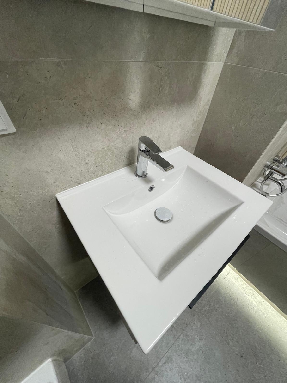 Biała, prostokątna umywalka z chromowaną baterią w nowoczesnej łazience z szarymi płytkami. Umywalka zamontowana na czarnej szafce z podświetleniem LED.