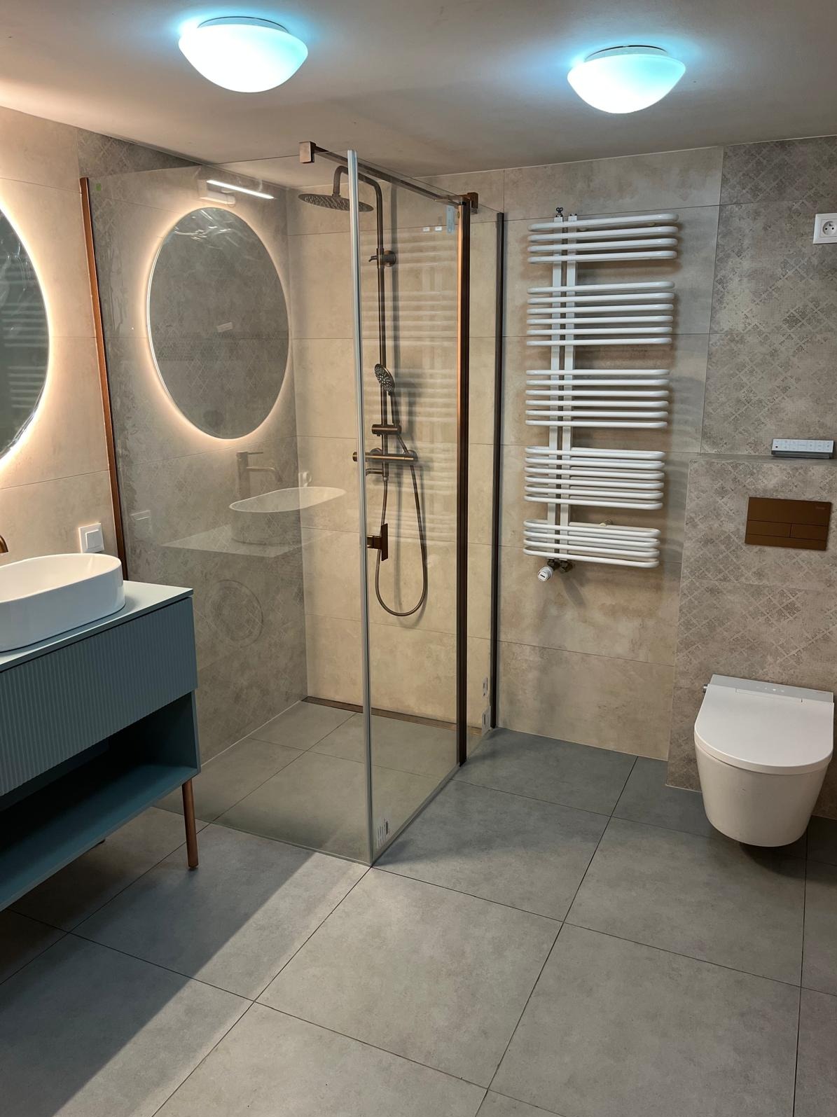 Nowoczesna łazienka z kabiną prysznicową typu walk-in, umywalką na szafce w odcieniach błękitu i podświetlanym lustrem. Szare płytki podłogowe i dekoracyjne na ścianach.