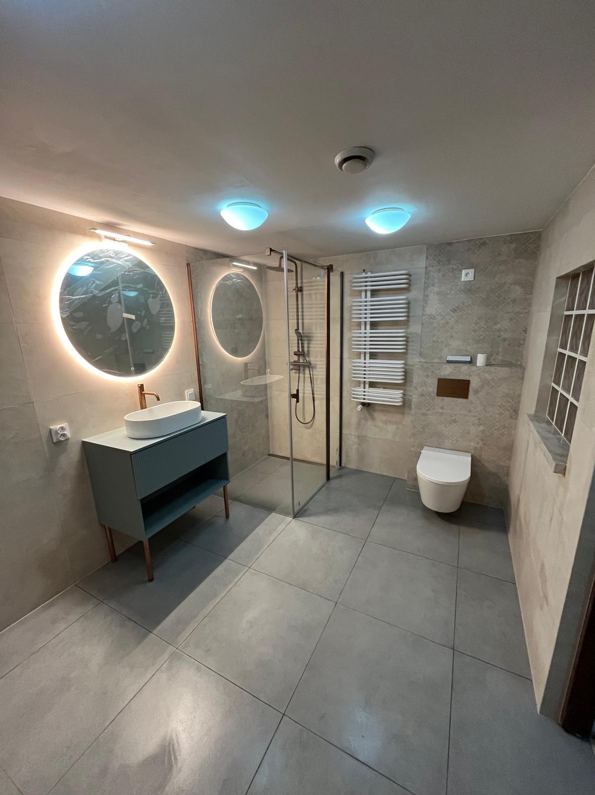 Wykończona łazienka w szarościach z okrągłymi lustrami z podświetleniem LED, kabiną prysznicową typu walk-in i grzejnikiem drabinkowym. Nowoczesny design.
