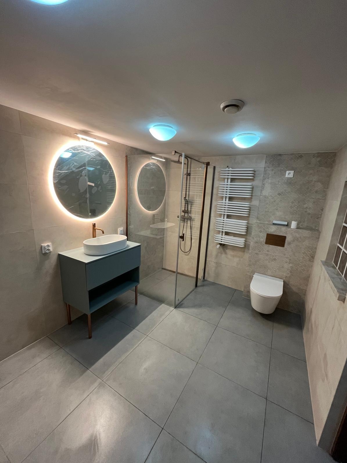 Nowoczesna łazienka z szarą armaturą, okrągłym lustrem z podświetleniem LED, kabiną prysznicową i grzejnikiem drabinkowym. Minimalistyczny design, płytki w odcieniach szarości.