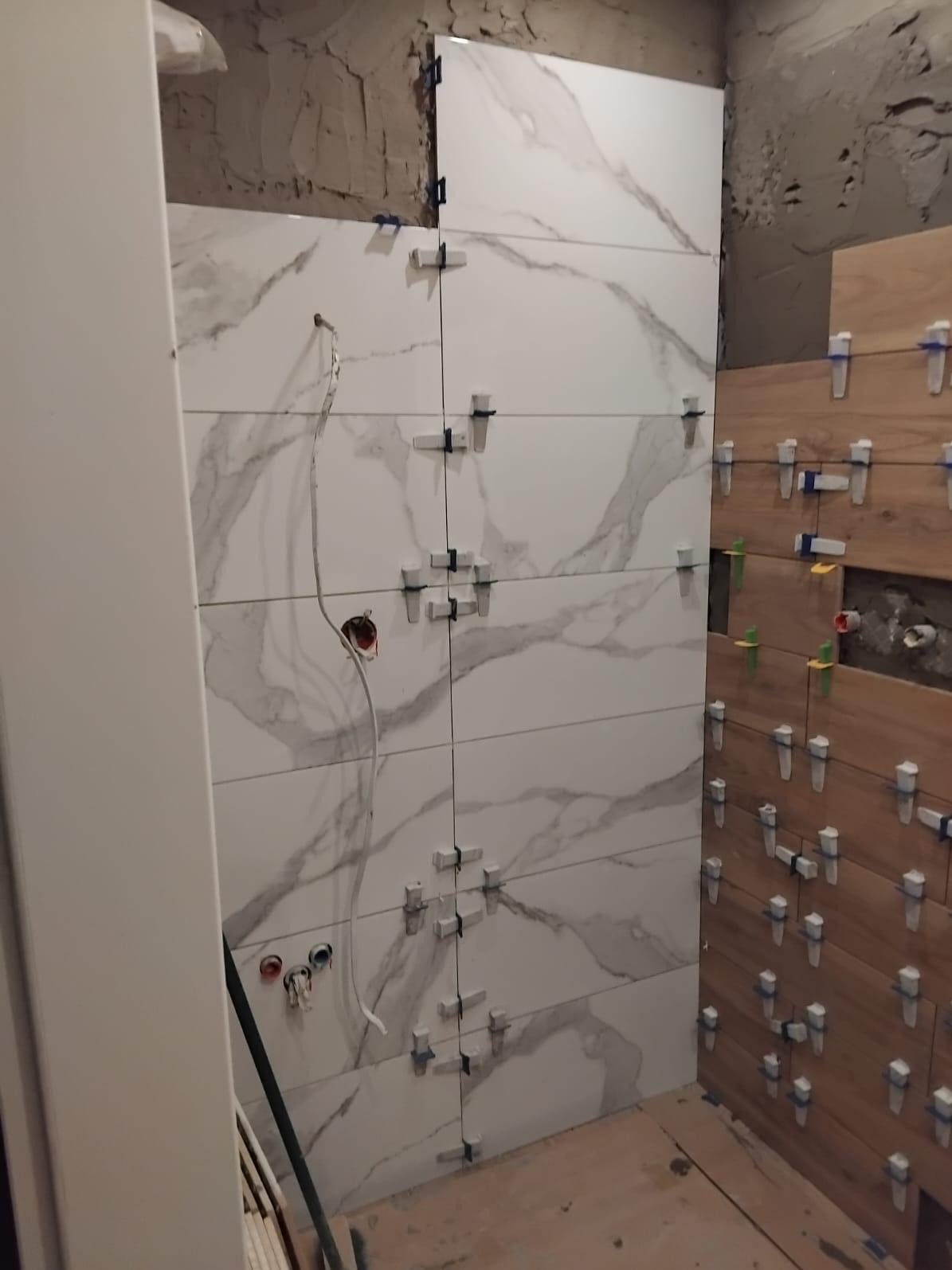Układanie płytek imitujących marmur na ścianie łazienki, z widocznymi krzyżykami dystansowymi i fragmentem instalacji elektrycznej. Częściowo ułożone płytki drewnopodobne.