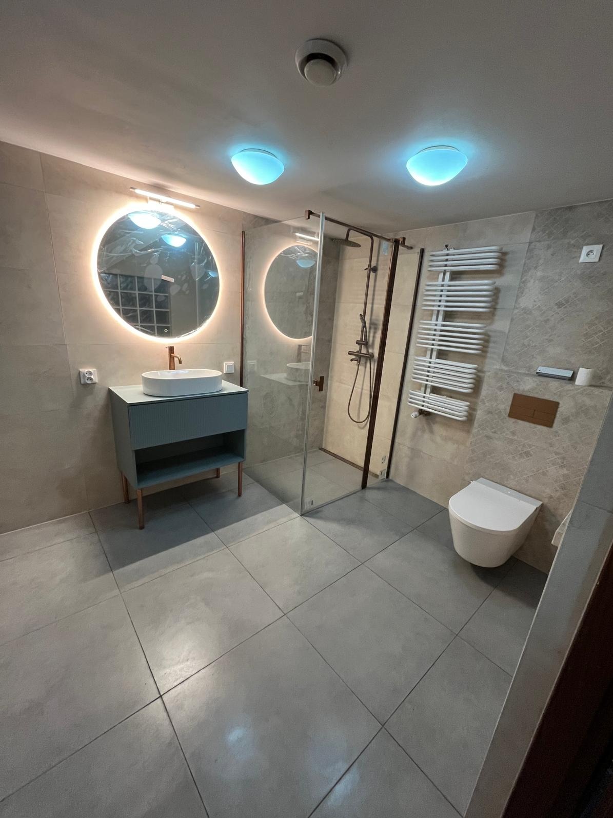 Nowoczesna łazienka z okrągłym lustrem podświetlanym LED, kabiną prysznicową typu walk-in i wiszącą toaletą. Szare płytki podłogowe i ścienne. Minimalistyczny design.
