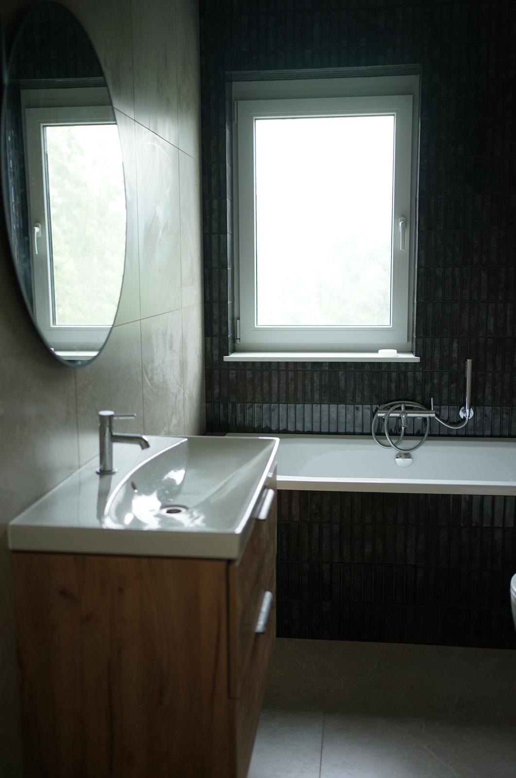 Nowoczesna łazienka z umywalką na szafce z drewnianym frontem, owalnym lustrem i wanną obłożoną ciemnymi, wąskimi płytkami. Okno z białymi ramami.