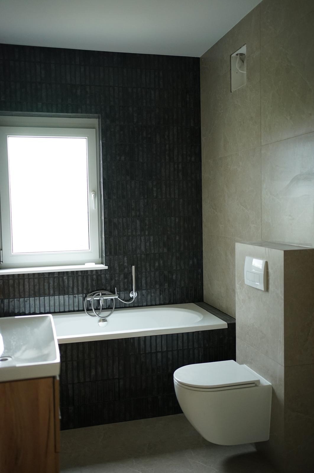 Nowoczesna łazienka z czarnymi płytkami imitującymi cegłę, białą wanną i podwieszaną toaletą. Minimalistyczny design, jasne okno, szare płytki na ścianie.