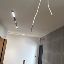 LuxSolutions - Nowoczesne wnętrze w trakcie prac elektrycznych: widoczne przewody wystające z sufitu, zamontowane listwy LED. Schody z drewnianą balustradą prowadzą w dół.