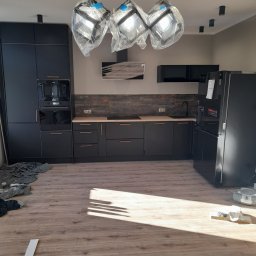 REMONTY WYKONCZENIA - Nowoczesna, czarna kuchnia z drewnianym blatem i podłogą. Lampy wiszące w folii ochronnej. Widoczne sprzęty AGD i elementy wykończeniowe.