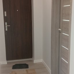 REMONTY WYKONCZENIA - Dwa rodzaje drzwi wewnętrznych: ciemne, pełne i jasne z przeszkleniami, zamontowane w jasnym korytarzu z panelami podłogowymi. Widoczna mata pod drzwiami.