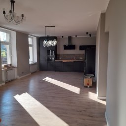 REMONTY WYKONCZENIA - Przestronne wnętrze po remoncie: parkiet, szara ściana, czarna zabudowa kuchenna z oświetleniem. Widoczne okna z naturalnym światłem i karton na podłodze.