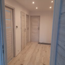 REMONTY WYKONCZENIA - Jasne, drewnopodobne panele podłogowe i trzy zamontowane drzwi wewnętrzne w korytarzu z oświetleniem punktowym. Widok zza otwartych drzwi.