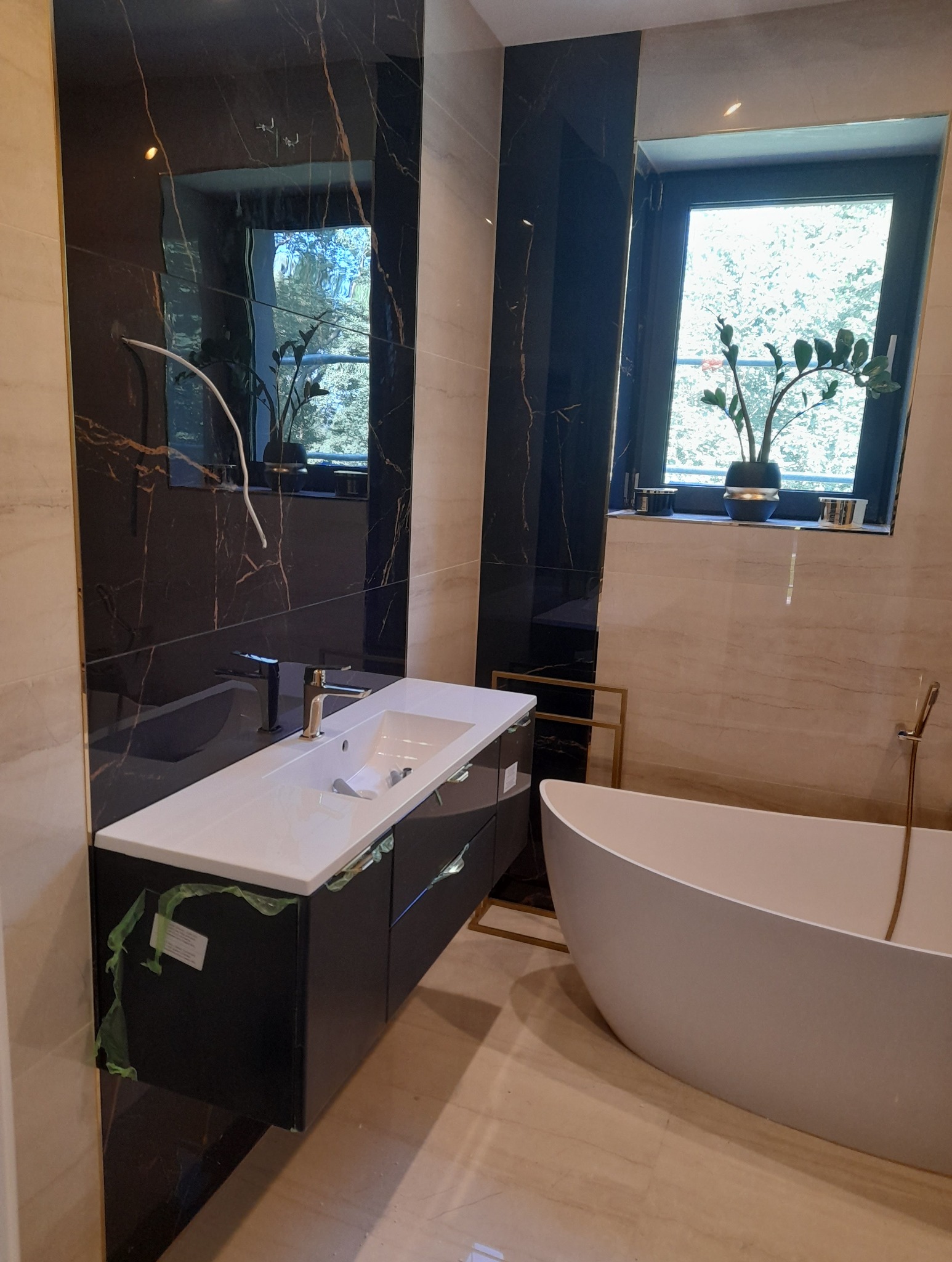 Nowoczesna łazienka z czarnymi płytami imitującymi marmur, białą umywalką z szafką i owalną wanną. Widoczne okno z rośliną doniczkową i złote detale.