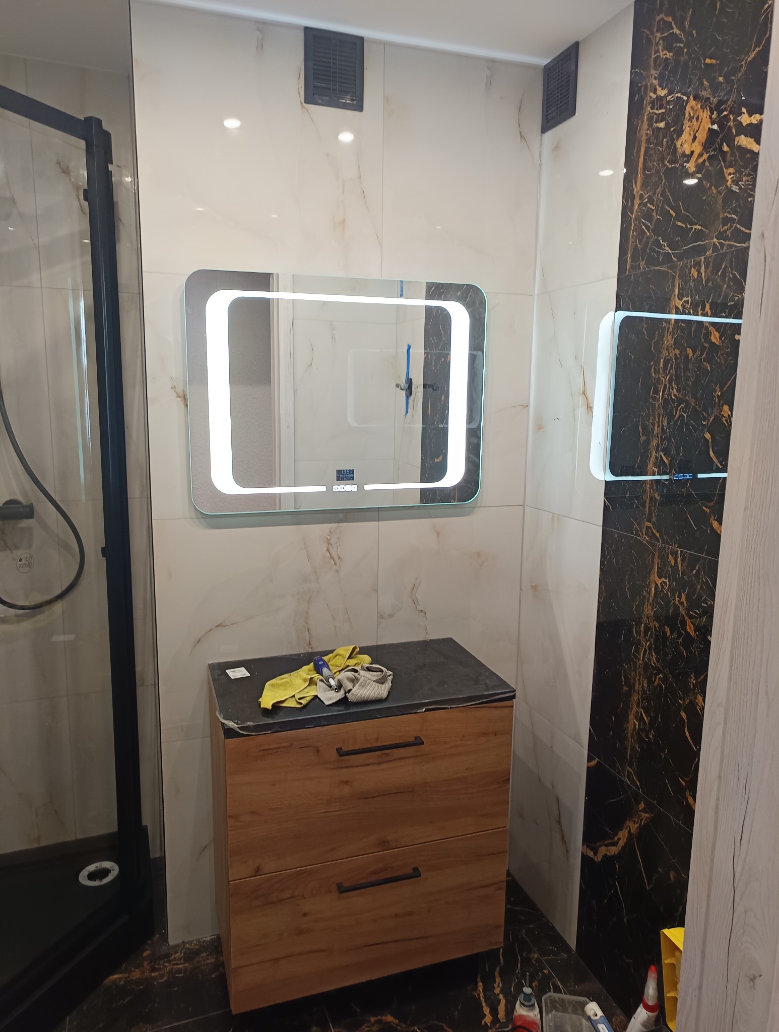 Wykończona łazienka z kabiną prysznicową, szafką z drewnianymi frontami i podświetlanym lustrem. Na blacie leżą narzędzia i ściereczka.