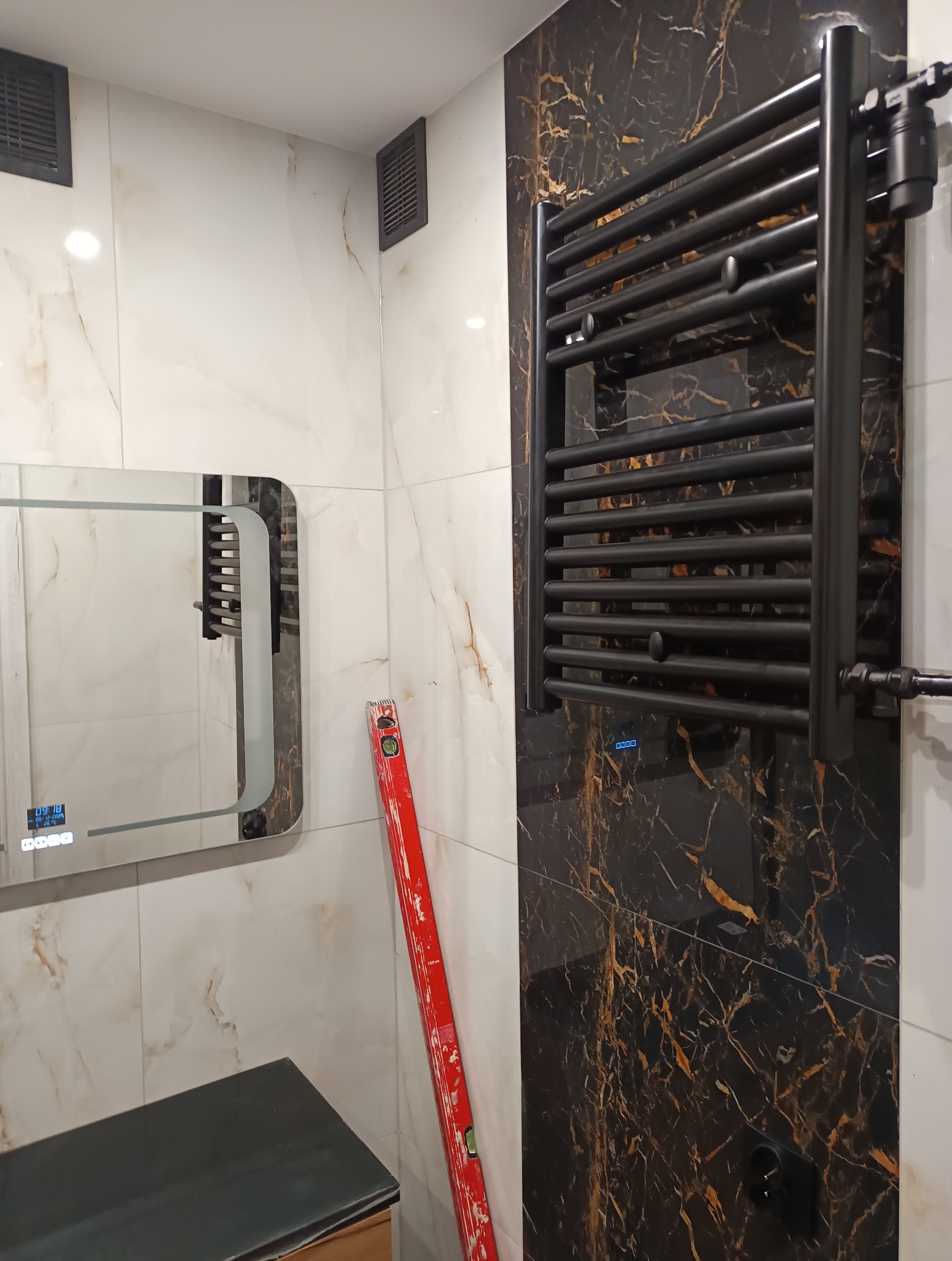 Nowoczesna łazienka z czarnym grzejnikiem drabinkowym na tle marmurowych płytek. Widoczny fragment lustra z podświetleniem LED i poziomica oparta o ścianę.