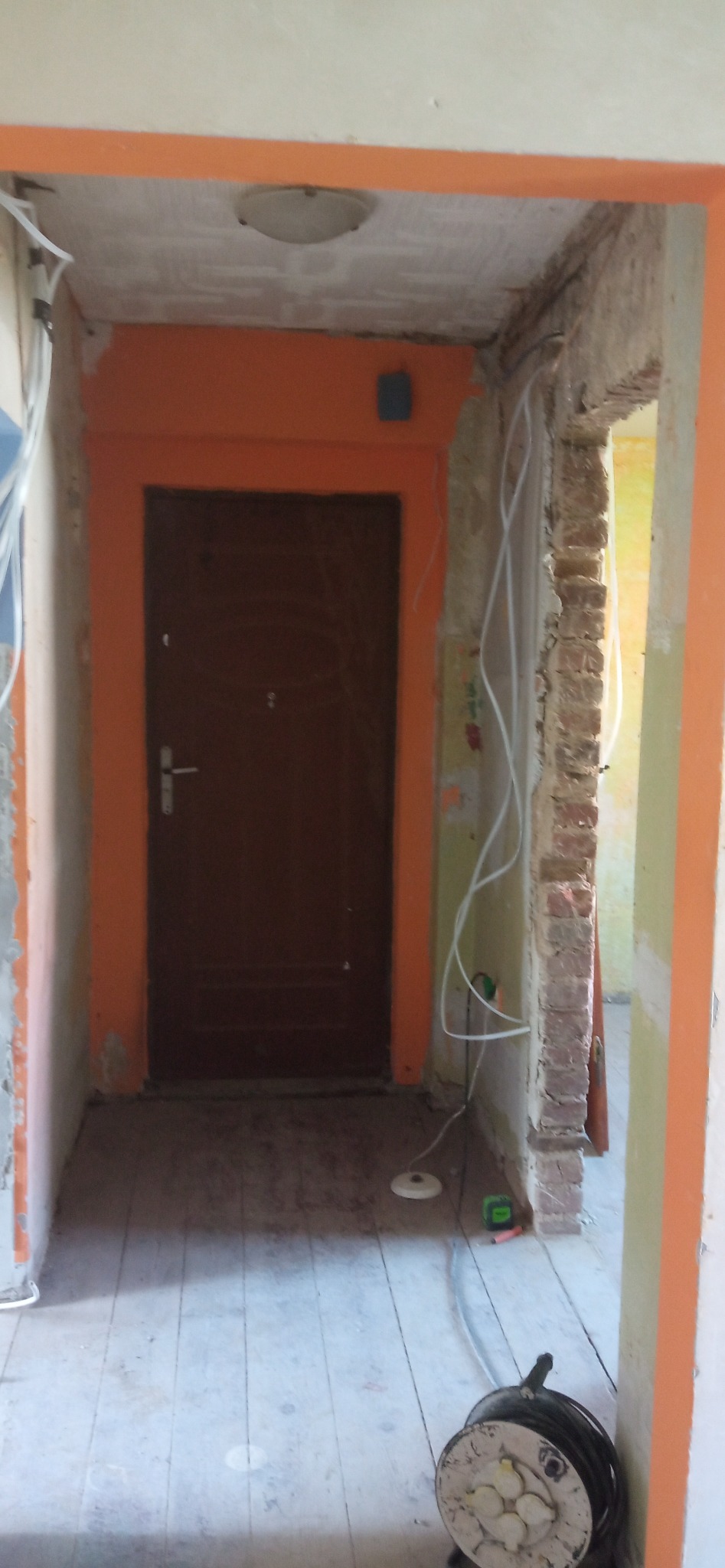Remont korytarza: brązowe drzwi z pomarańczową ościeżnicą, odsłonięta cegła, kable elektryczne i zwinięty przedłużacz na drewnianej podłodze. Widoczne ślady prac remontowych.