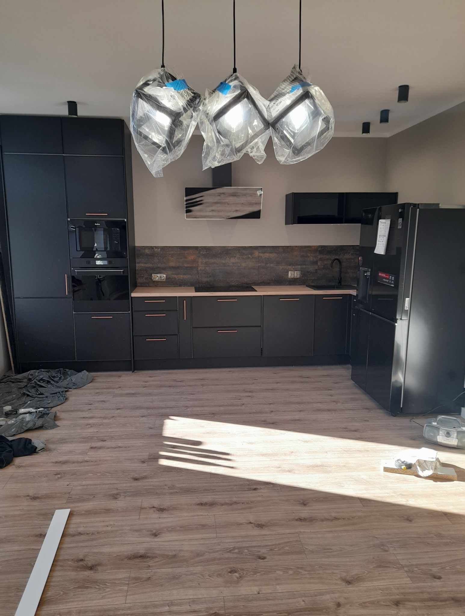 Nowoczesna, czarna kuchnia z drewnianym blatem i podłogą. Lampy wiszące w folii ochronnej. Widoczne sprzęty AGD i elementy wykończeniowe.