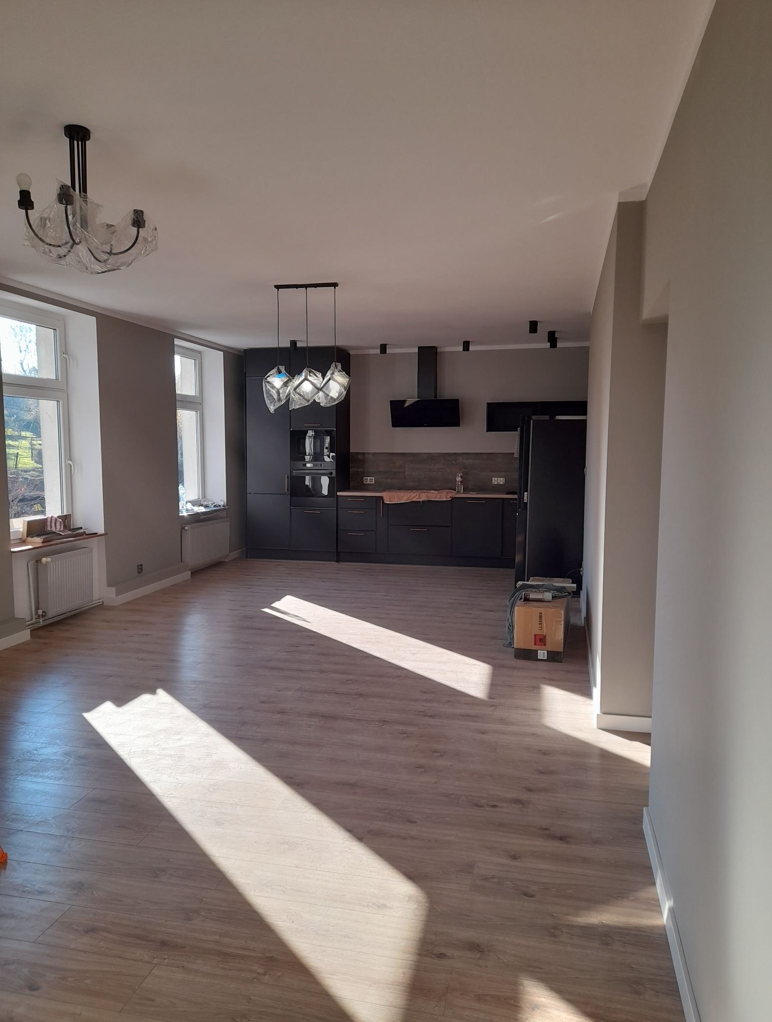 Przestronne wnętrze po remoncie: parkiet, szara ściana, czarna zabudowa kuchenna z oświetleniem. Widoczne okna z naturalnym światłem i karton na podłodze.