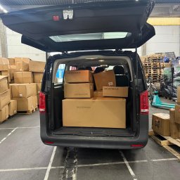 Northumberland GmbH - Ciemny van z otwartym bagażnikiem, wypełniony kartonami z logo 'VA VITOMALIA', na tle magazynu z paletami i kartonami. Widoczne ślady opon na podłodze.