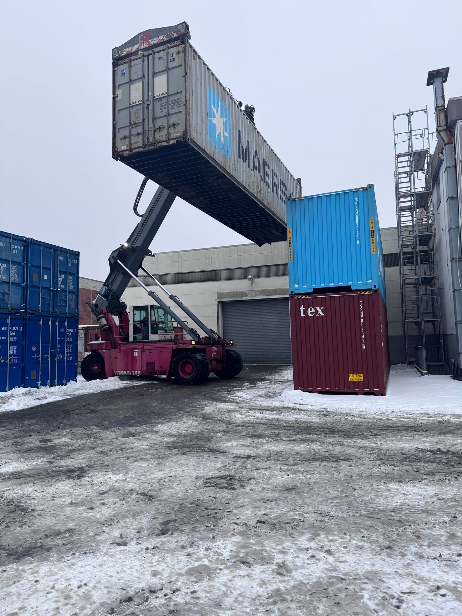 Wózek widłowy Ferrari 258 podnosi kontener Maersk na placu składowym. W tle budynek magazynowy i ustawione kontenery. Zimowa sceneria z resztkami śniegu.