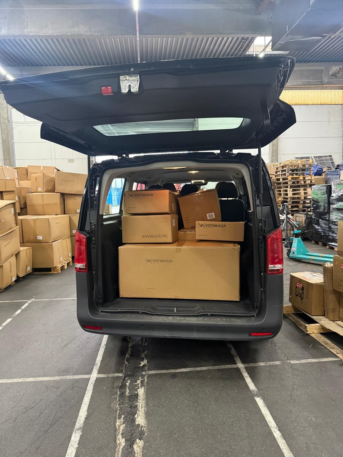 Ciemny van z otwartym bagażnikiem, wypełniony kartonami z logo 'VA VITOMALIA', na tle magazynu z paletami i kartonami. Widoczne ślady opon na podłodze.