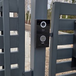 Blue-MonterPRO Patryk Gralak - Nowoczesny wideodomofon Kenik zamontowany na grafitowej bramie posesji. Widoczne drzewa w tle. Minimalistyczny design urządzenia.