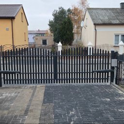 Blue-MonterPRO Patryk Gralak - Nowoczesna, czarna brama automatyczna z napędem ramieniowym, zamontowana przed domem z kostki brukowej. Widoczne dwa budynki w tle, jeden w odcieniach żółci, drugi beżowy.