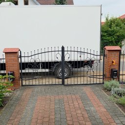 Blue-MonterPRO Patryk Gralak - Kuta brama wjazdowa z automatyką, czarna, z dekoracyjnymi elementami, zamontowana na ceglanych słupkach. W tle biały samochód dostawczy i dom z brązowym dachem.