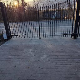 Blue-MonterPRO Patryk Gralak - Otwarta, metalowa brama posesji z automatyką, widok z zewnątrz na ulicę i zachodzące słońce. Kostka brukowa na pierwszym planie.