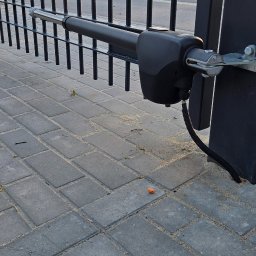 Blue-MonterPRO Patryk Gralak - Zamontowany napęd bramy skrzydłowej, widoczny na tle ogrodzenia i kostki brukowej. Element automatyki bram w zbliżeniu, z widocznym ramieniem i kablem.