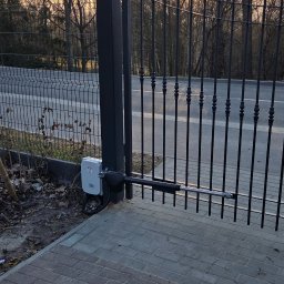 Blue-MonterPRO Patryk Gralak - Czarna brama z automatyką na tle drzew i nieba o zmierzchu. Widoczny siłownik i skrzynka sterownicza. Kostka brukowa na pierwszym planie.